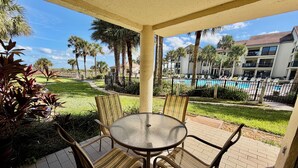 Eine überdachte Terrasse mit rundem Tisch und Stühlen, mit Blick auf einen Pool und eine angelegte Grünfläche mit Palmen.