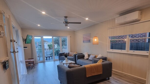 A spacious living area with a grey sofa, a small round table, and a ceiling fan. There is a TV mounted on the wall and a window with a view of the outside. - 5 min walk to the Beach! (Rosebud)