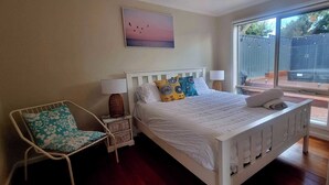 A bedroom with a white bed, a chair with floral upholstery, a bedside table with a lamp, and a window with a view of a fenced area. - 5 min walk to the Beach! (Rosebud)