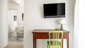 A room with a wooden desk, a green chair, a television mounted on the wall, and a lamp on the desk.