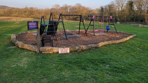 Ein Spielplatz mit Schaukeln und einer Rutsche, umgeben von einer Wiese und Bäumen.