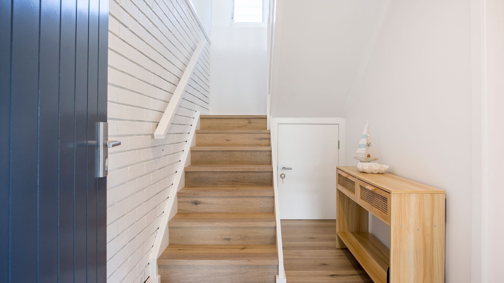 Un escalier avec des marches en bois et une rampe blanche, menant à une porte à côté de laquelle se trouve une petite console en bois.