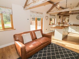 A cozy living room with a brown leather sofa, a black and white patterned rug, wooden beams on the ceiling, and a kitchen area with a wooden countertop and white cabinets.