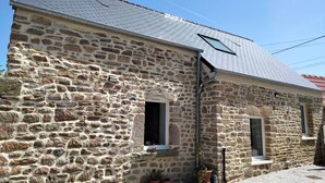 A stone building with a sloped roof, two windows, and a small porch area with potted plants. - House (Maupertus-sur-Mer)