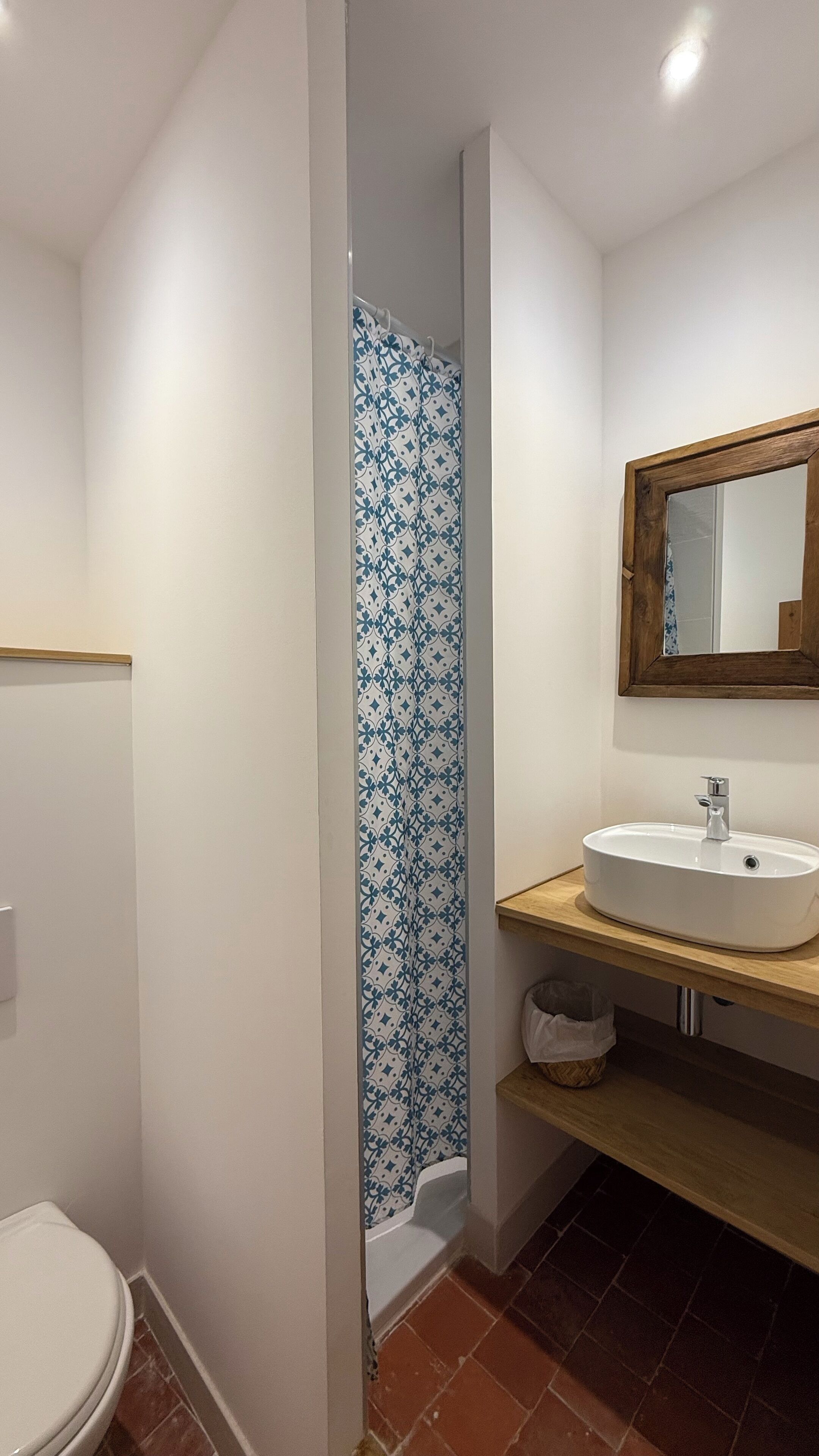 A bathroom with a white toilet, a shower with a blue and white patterned curtain, a wooden vanity with a white sink, and a mirror with a wooden frame.
