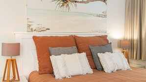 Quarto com cama, duas luminárias de cabeceira, encosto e uma pintura com tema de praia acima da cama.