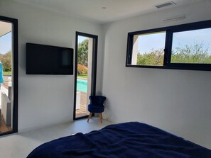 Quarto com cama, televisão montada na parede, uma cadeira e vista para uma piscina através de uma porta de vidro.