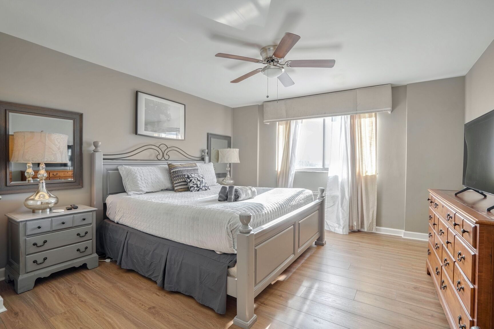 Un dormitorio con cama, mesitas de noche, cómoda, ventilador de techo y ventana con cortinas.