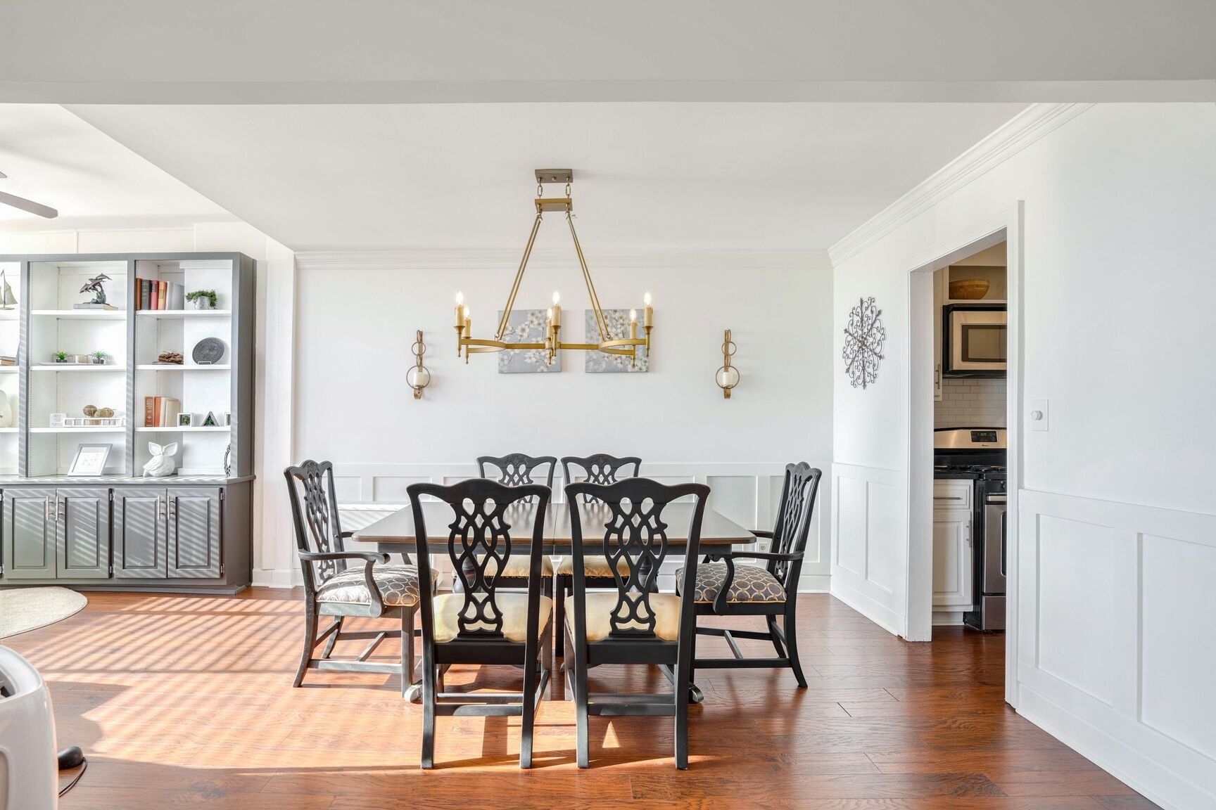 A dining area with a wooden table and chairs, a chandelier, and a white cabinet with shelves.