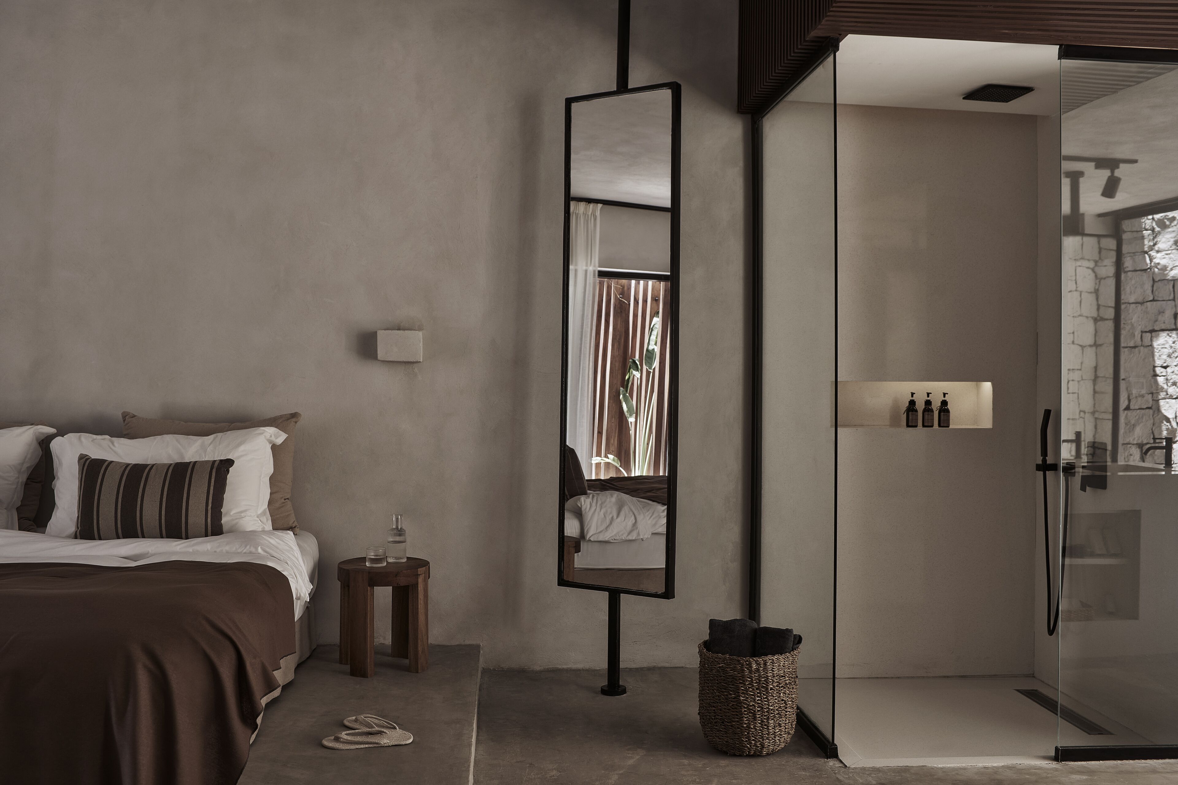A modern bedroom with a large bed, a bedside table with a lamp, a mirror, and a glass-enclosed shower area.