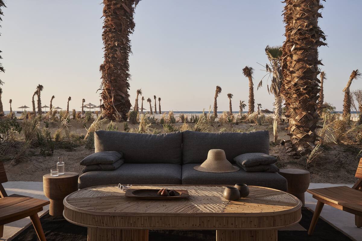 A beachside lounge with a sofa, round table, and wooden chairs. Palm trees and the ocean are visible in the background.