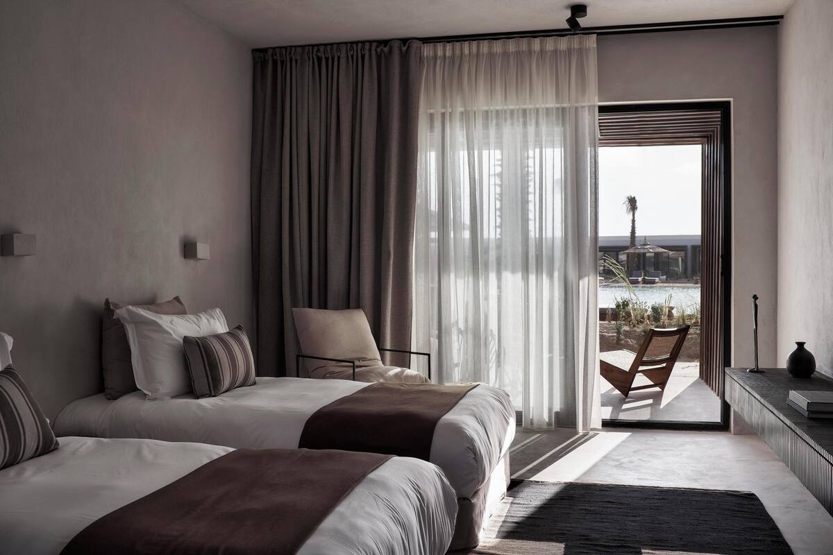 A hotel room with two beds, a chair, a view through a glass door, and a small table with a vase.