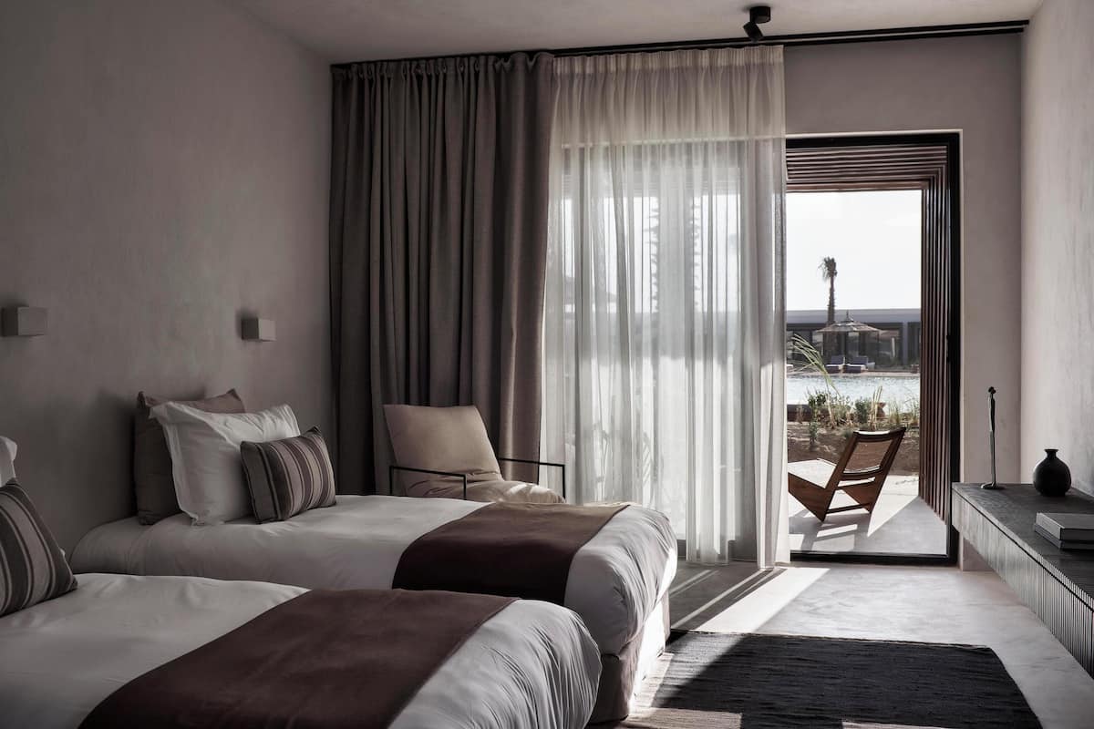 A hotel room with two beds, a chair, a view through a glass door, and a small table with a vase.