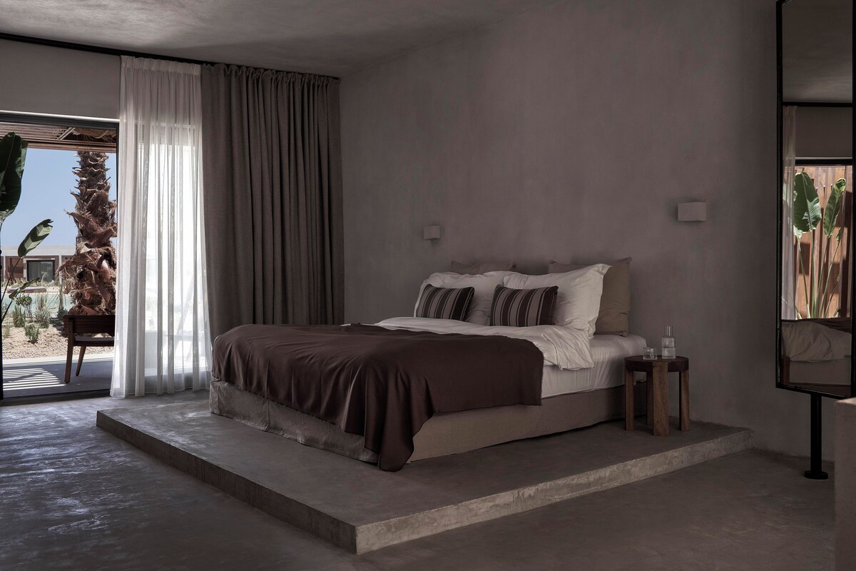 A modern bedroom with a bed, bedside tables, and a view of a palm tree outside.