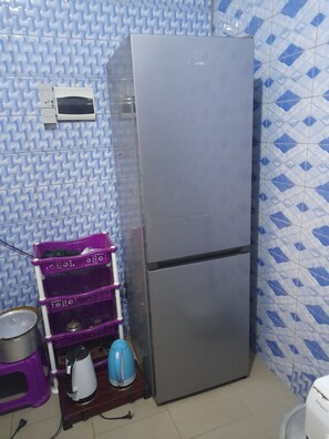 A compact kitchen with a stainless steel refrigerator, a purple and white laundry basket, a kettle, and a small sink.