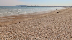 A pebbled beach with calm waters, a distant shoreline, and a few people in the distance. - Beach Retreat (Weymouth)