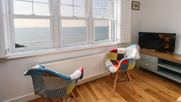 A room with a large window overlooking the sea, a television on a wooden stand, and two colorful chairs. - Beach Retreat (Weymouth)