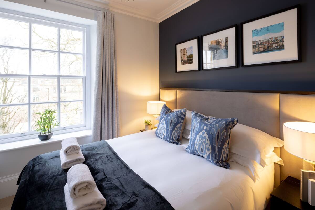 A hotel room with a large bed, bedside lamps, a window with a view, and framed pictures on the wall.