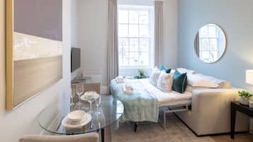 A modern hotel room with a glass dining table, a bed with a green throw, a round mirror, and a view of the city through the window.