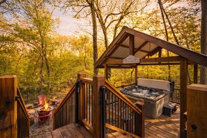 A wooden deck with a hot tub and a fire pit, surrounded by trees. - Seasonal Creek • Hot Tub • Playground • Game Loft (Broken Bow)