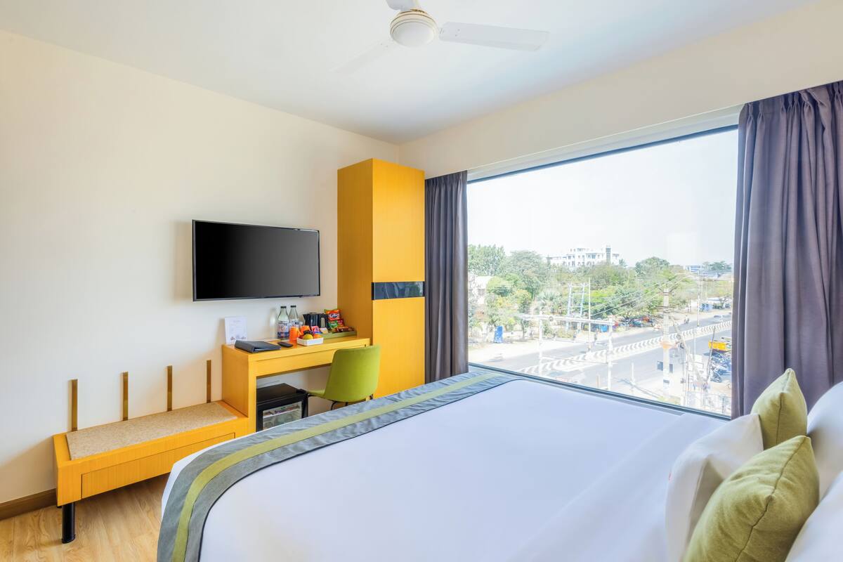 Habitación de hotel con cama, escritorio, silla, televisor y un ventanal con cortinas.