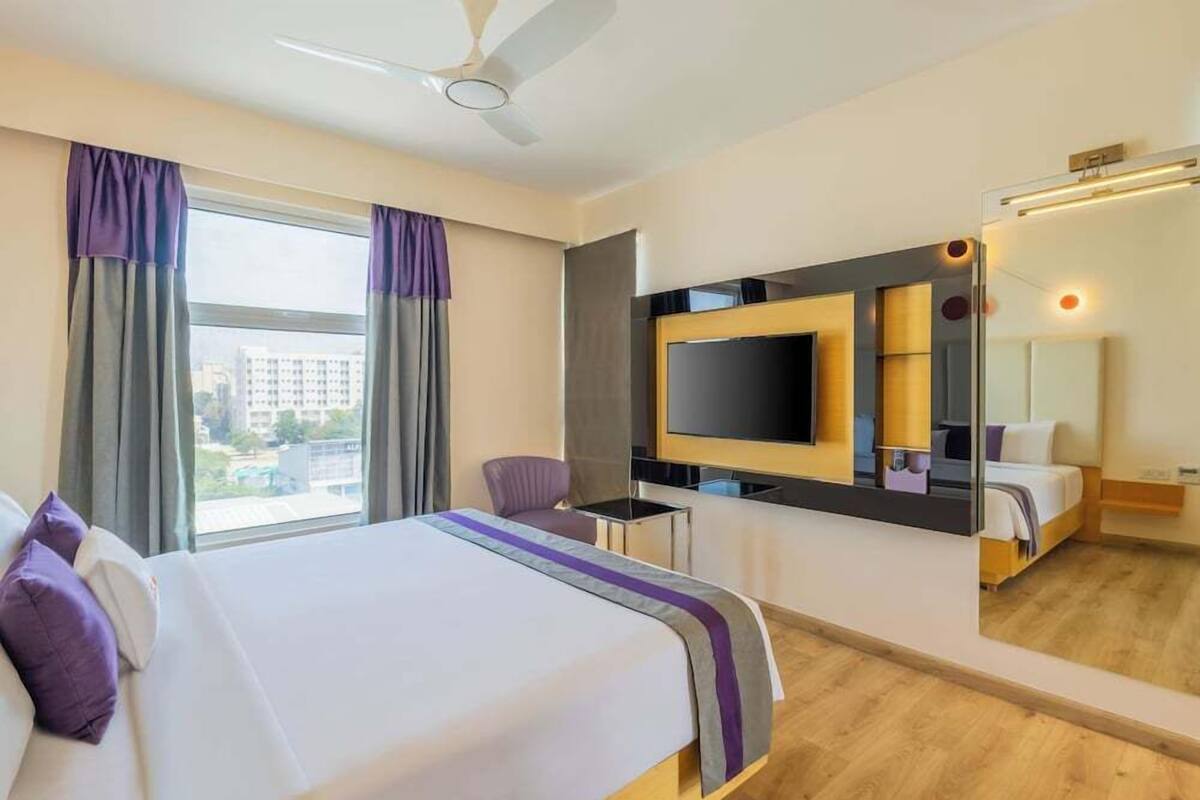 Una habitación de hotel con cama, televisión, espejo y ventana con cortinas.