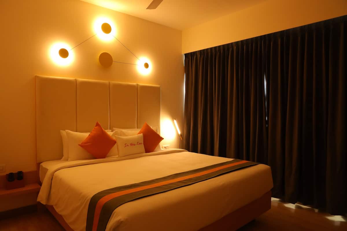 Una habitación de hotel con una cama grande, cabecera con luces circulares, mesita de noche con dos tazas y una ventana con cortinas oscuras.