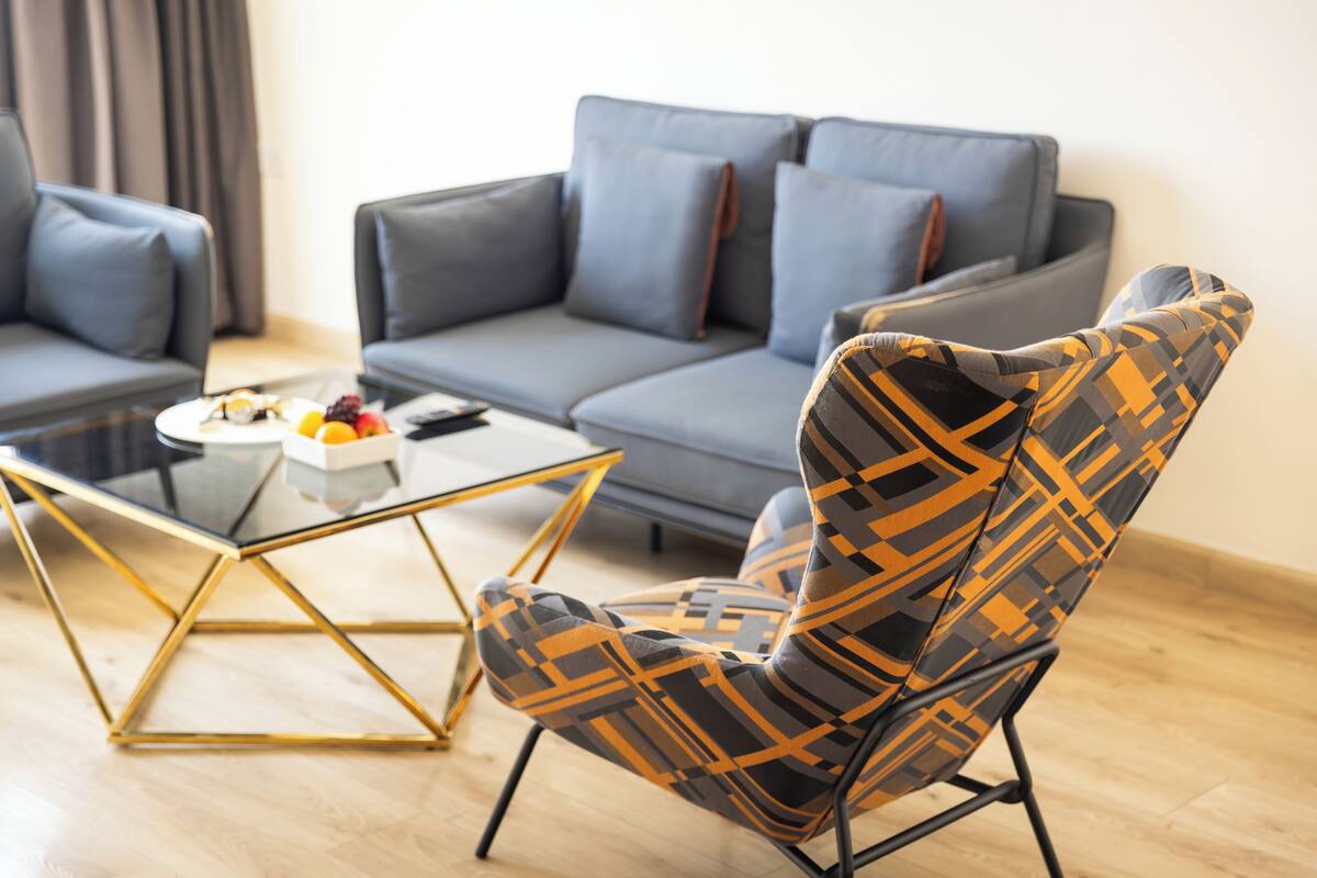 Una sala de estar moderna con un sofá gris, un sillón estampado y una mesa de centro de vidrio con fruta.
