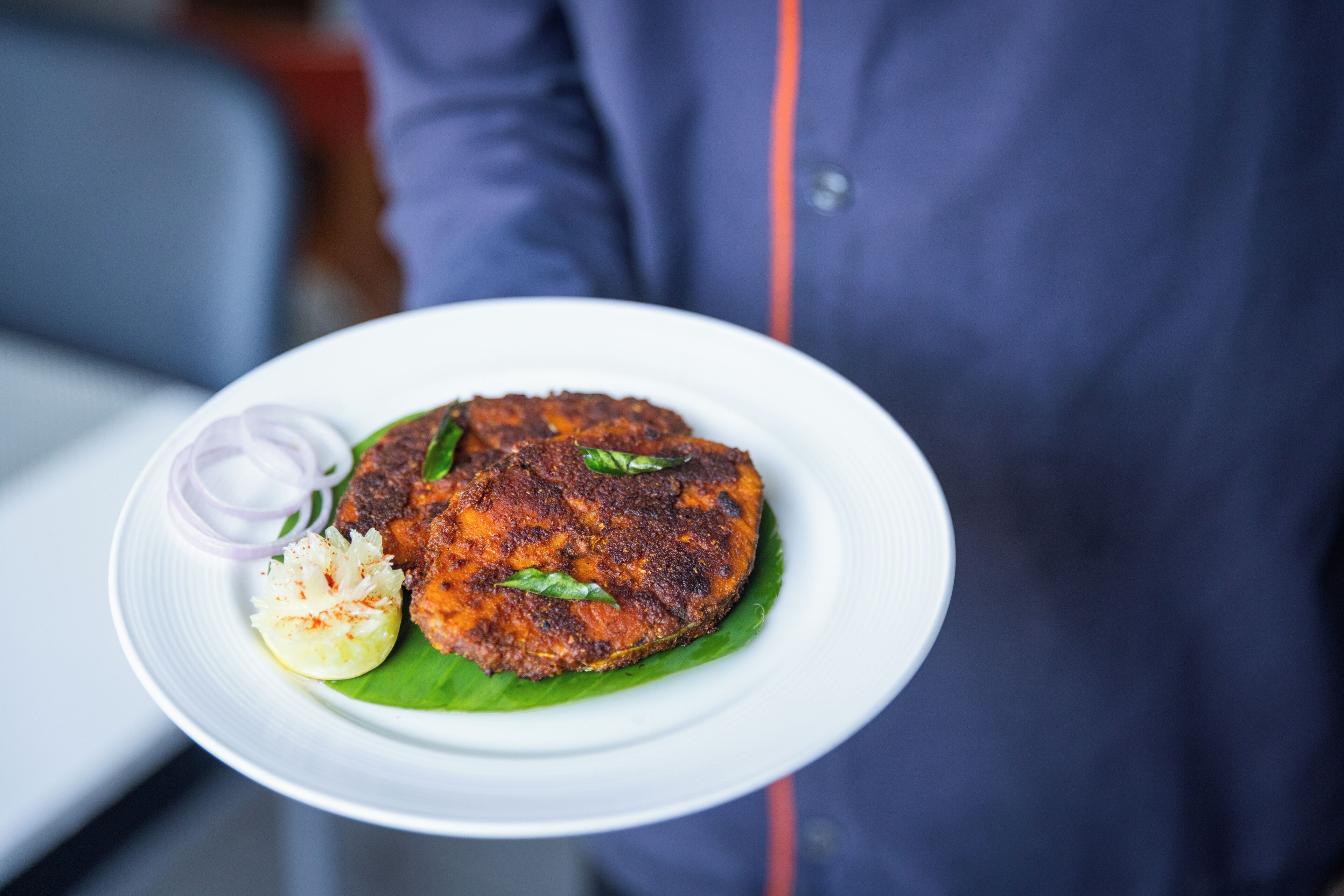 A plate with a food item, possibly a fried patty, garnished with sliced onions and a wedge of lemon, served on a white plate with a green leaf underneath.