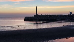 Paisaje costero con un faro, mar tranquilo y una playa arenosa.