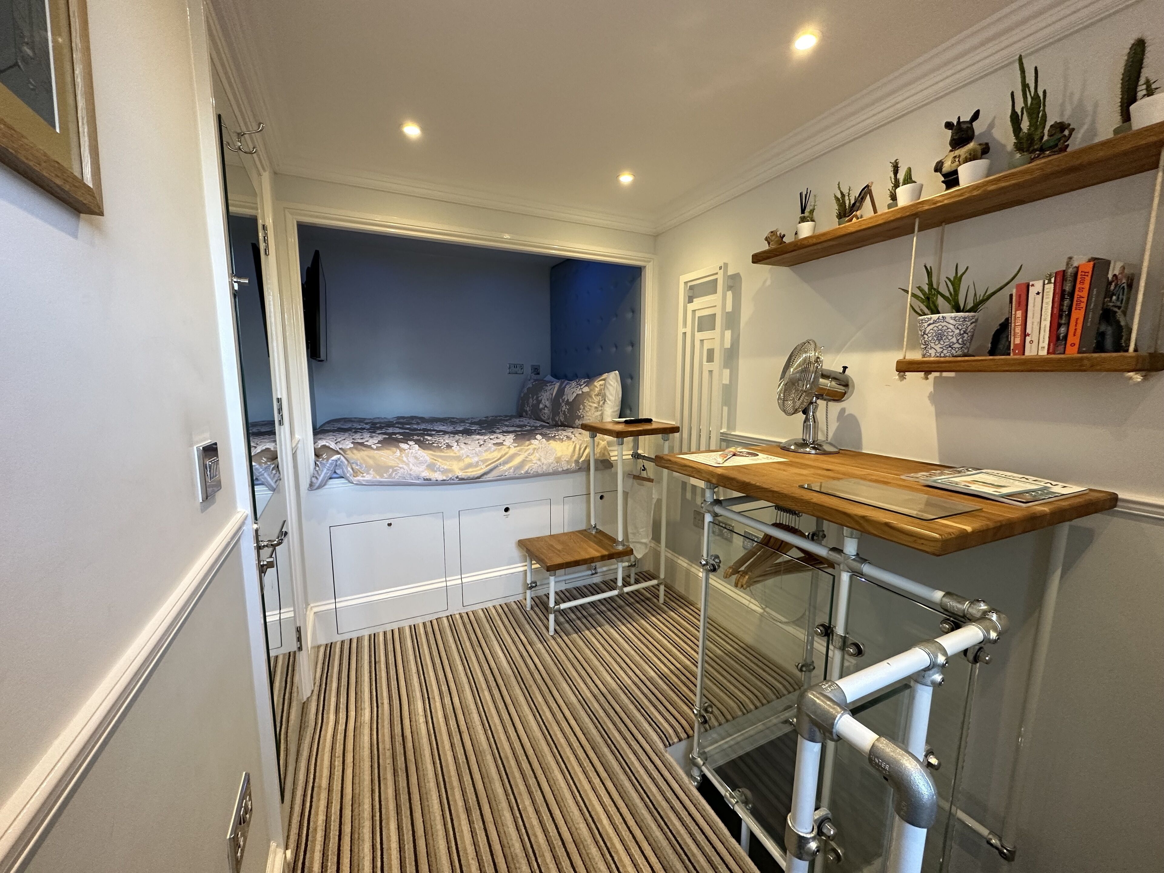 Ein gemütliches Schlafzimmer mit Bett, Nachttisch und einem kleinen Hocker. Ein Regal zeigt Pflanzen und Bücher, und ein Ventilator steht auf einem Gestell.