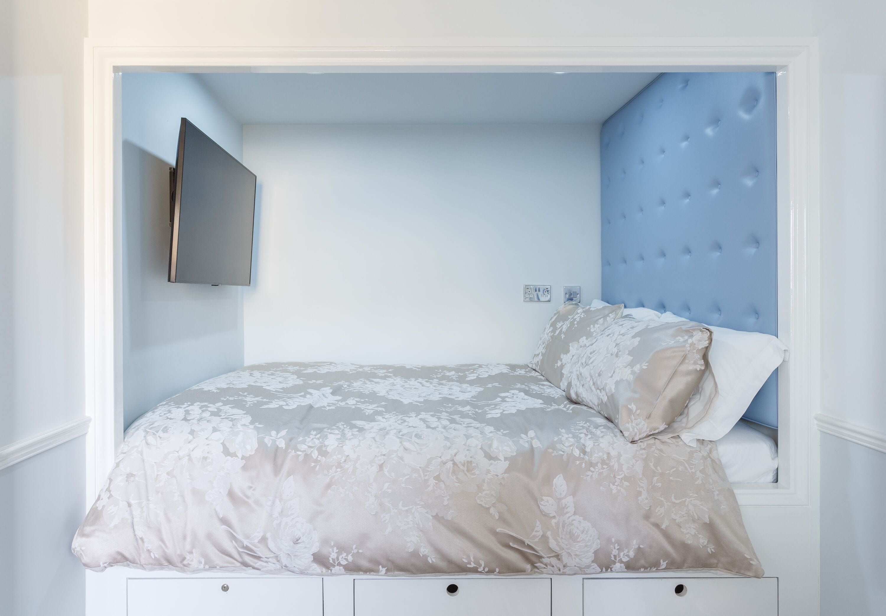 A bedroom with a bed, a wall-mounted TV, and a window with a blue headboard.