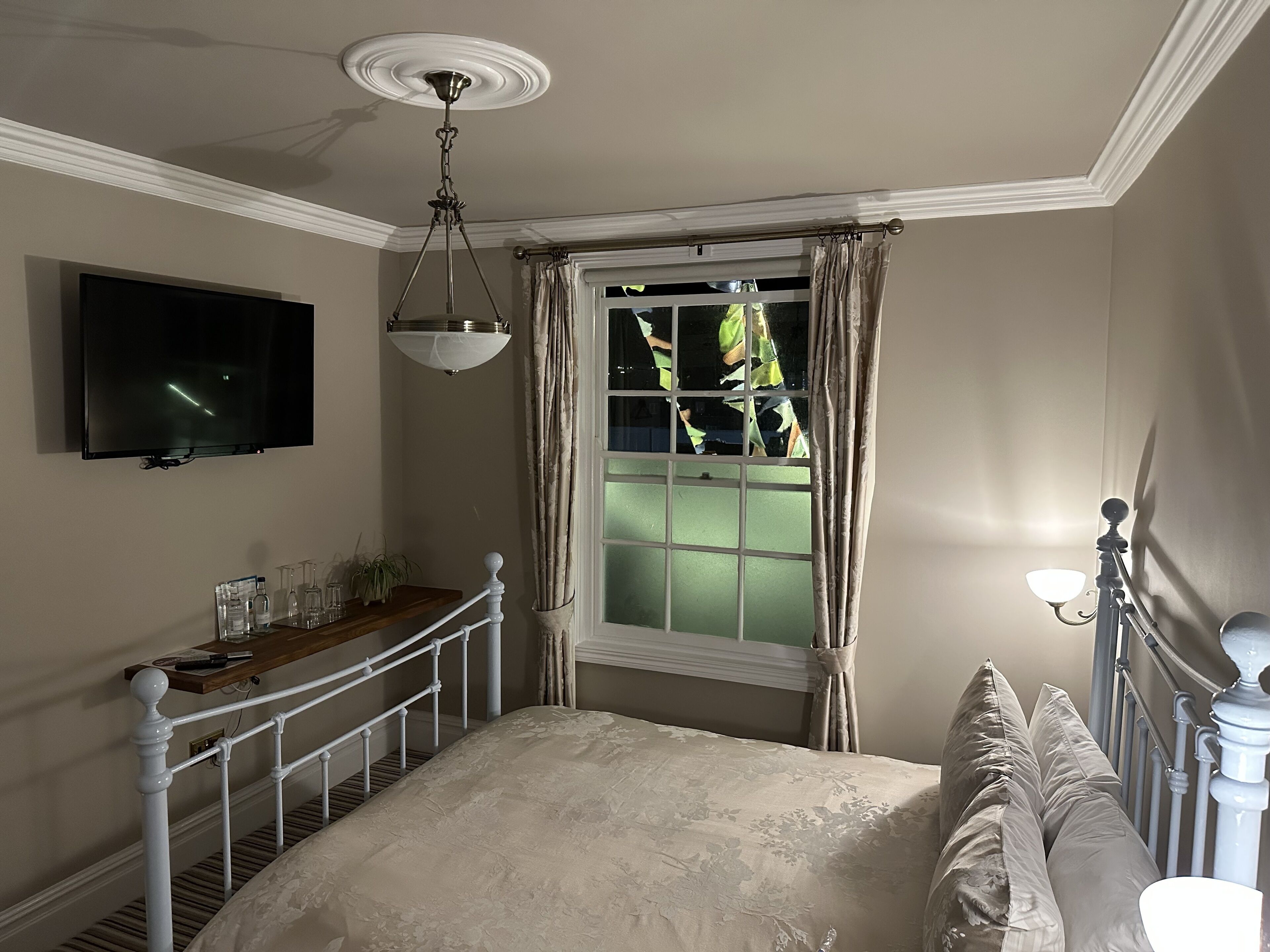 A bedroom with a bed, a TV mounted on the wall, a window with curtains, and a bedside table with a lamp.
