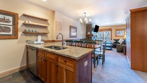 A kitchen with granite countertops, wooden cabinets, and a built-in dishwasher. There is a dining area with a table and chairs, and a living room with a sofa and a TV. - Ideally Located Condo w/ Shared Pool & Hot Tub and Private Balcony w/ Views! (Keystone)