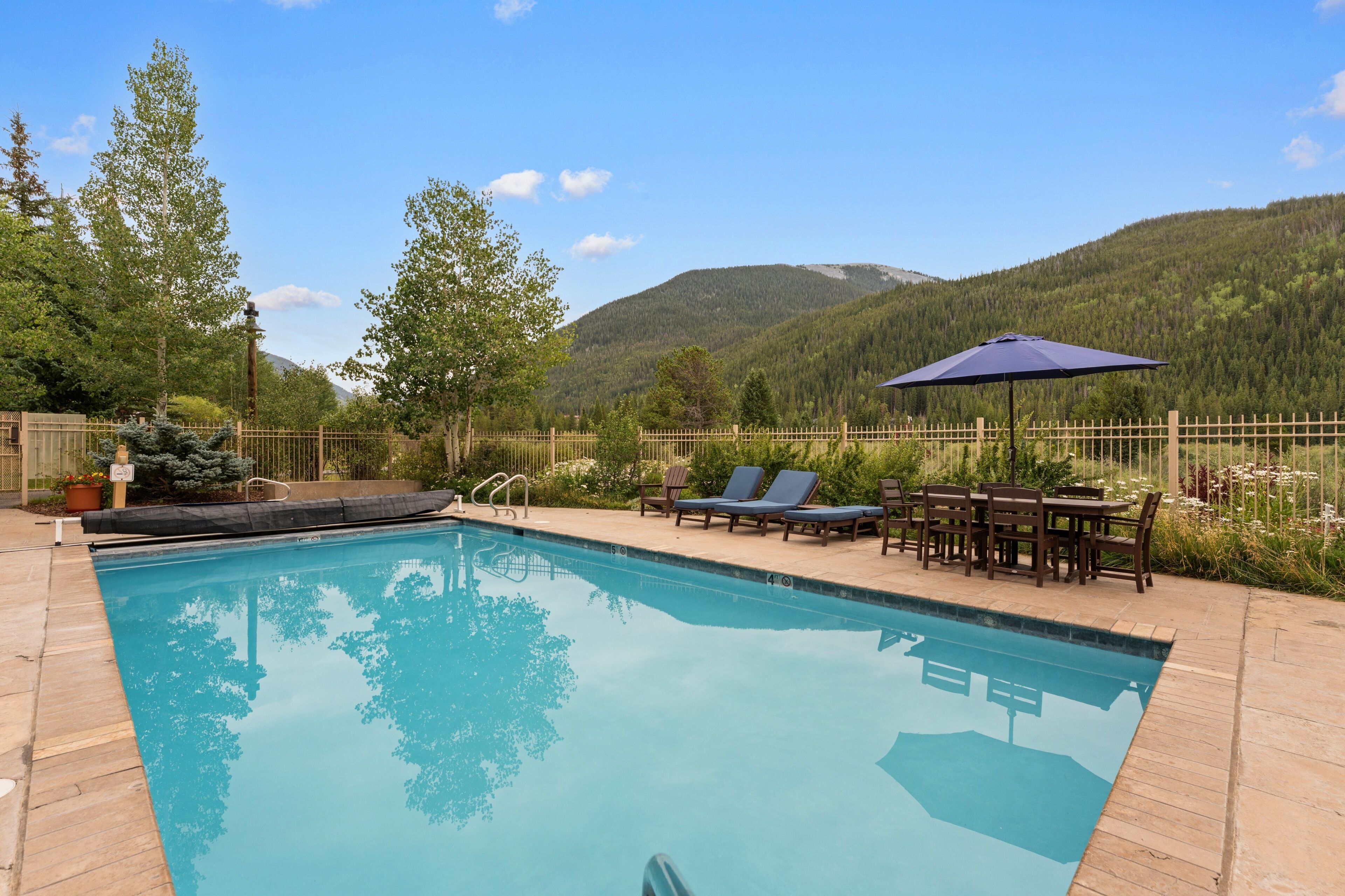 A swimming pool with lounge chairs and a table, surrounded by a wooden fence and trees, with mountains in the background.