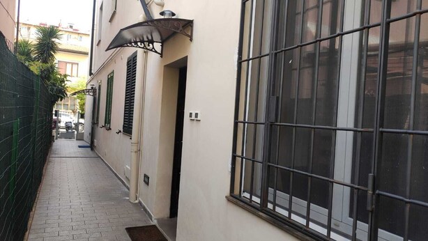 A narrow alleyway with a building featuring a large window with metal bars, a door, and a small outdoor light fixture. - VACATION HOME RICKY-LERUA (Firenze)