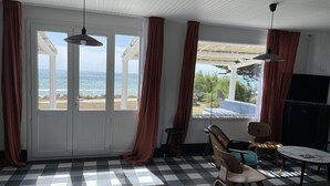 A room with a checkered floor, a TV, a table, and chairs. There are windows with curtains, and a view of the beach and ocean.