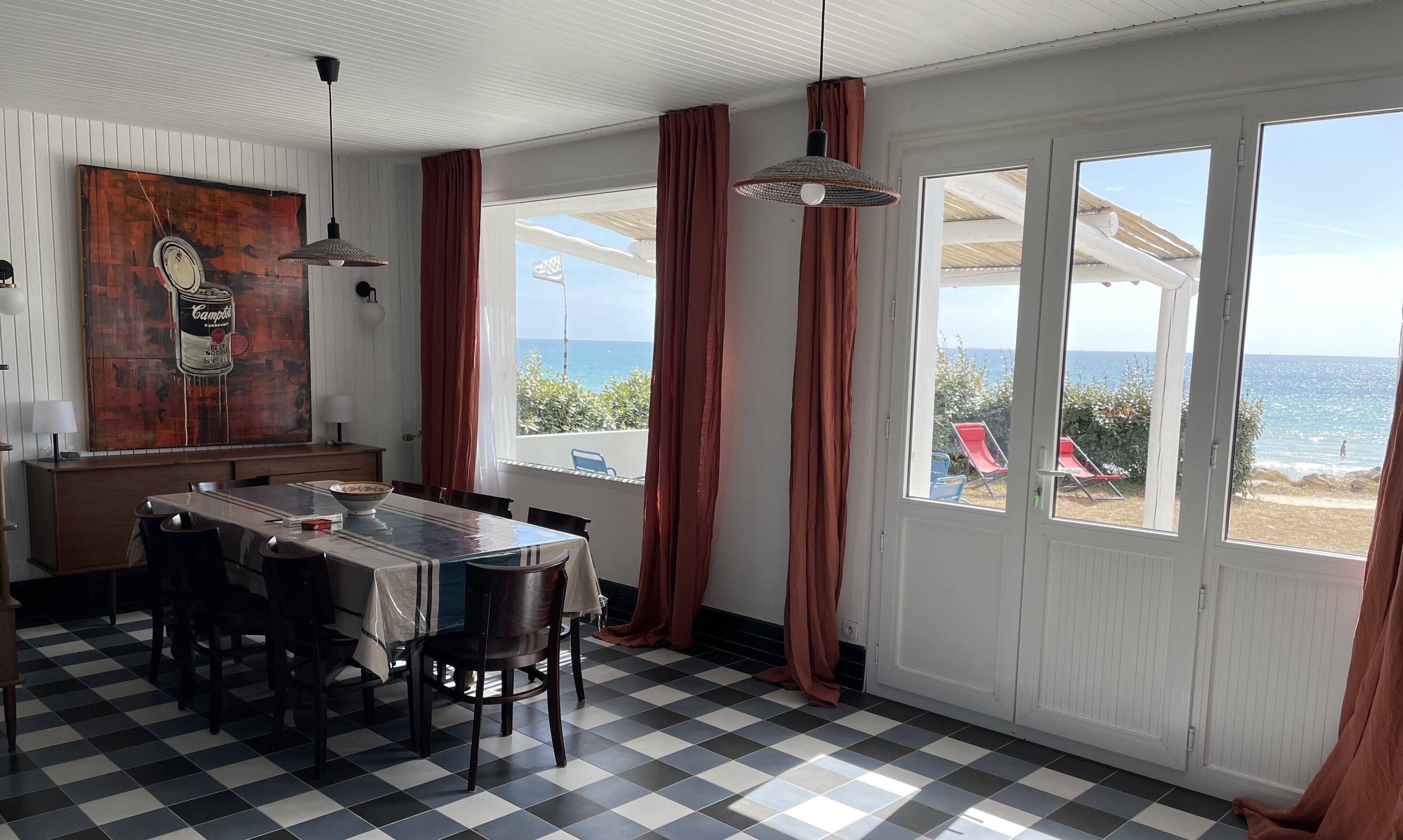 Un comedor con suelo a cuadros, una mesa con sillas, un cuadro en la pared y ventanas grandes con cortinas.