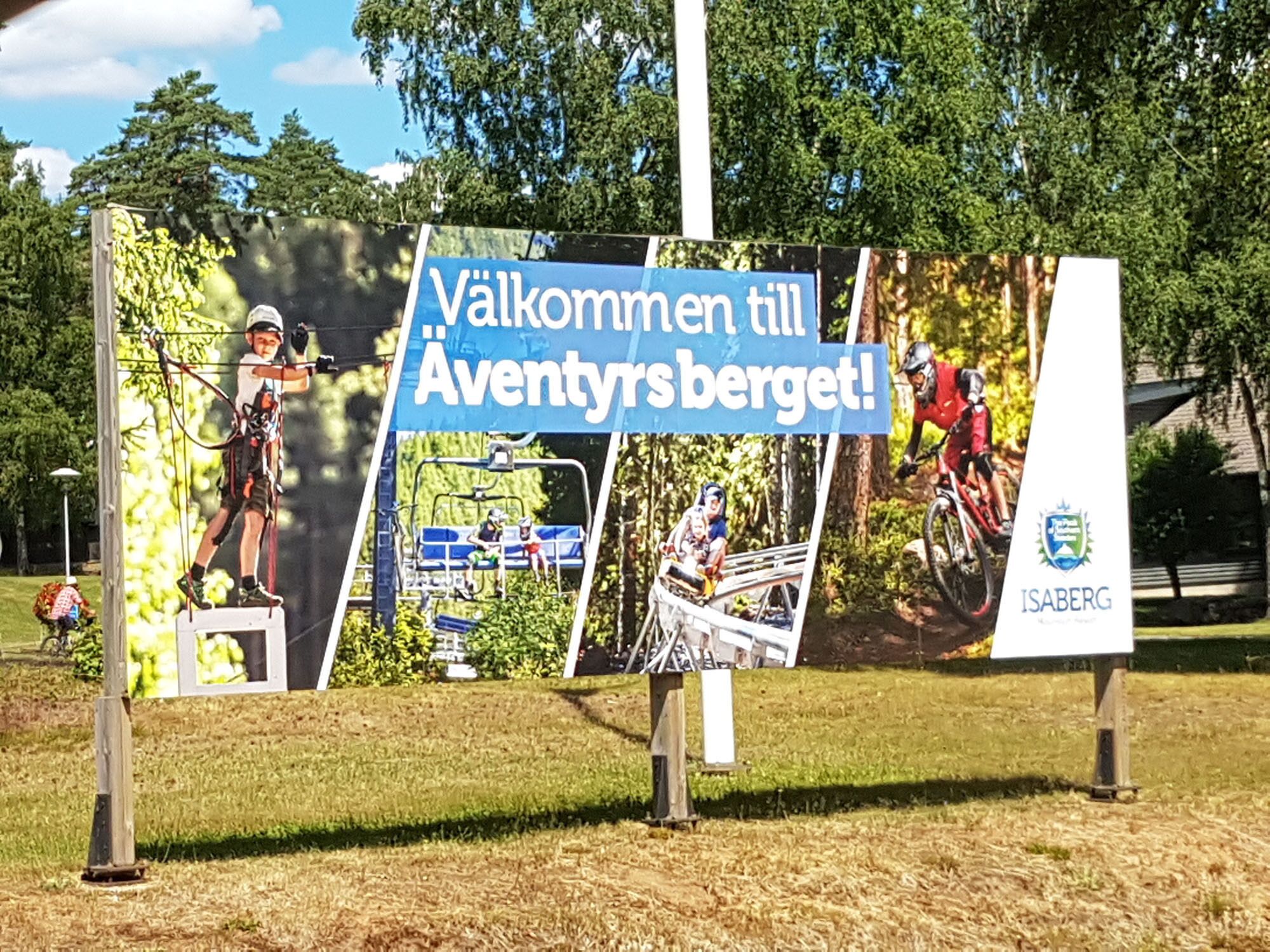 A sign with outdoor activities, including archery, a ski lift, and mountain biking, with the text "Välkommen till Adventysbergen!" and the logo "ISABERG".