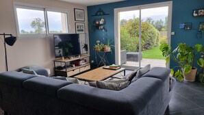 A living room with a sofa, a coffee table, a TV, and a bookshelf. There is a window and a door leading to a garden. - Spacious and quiet house (LA FRESNAIS)