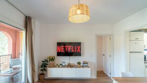 A modern living room with a large TV mounted on the wall, a wooden dining table, and a cozy seating area with a balcony view.
