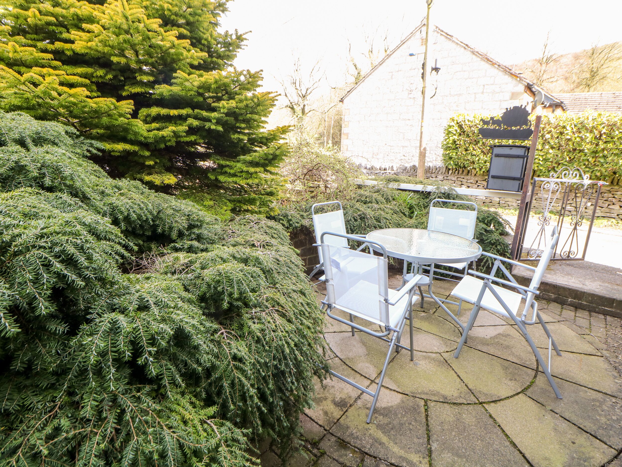 Een terras met een ronde tafel en vier stoelen, omgeven door weelderige begroeiing en een stenen muur.