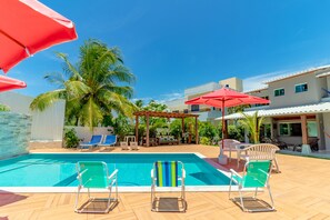 A swimming pool with lounge chairs and umbrellas, surrounded by palm trees and a covered seating area. -  GB60 Excellent House 4 Suites (Camaçari)