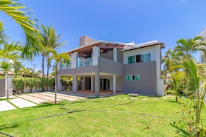 Uma casa moderna de dois andares com varanda coberta, cercada por vegetação exuberante e palmeiras.