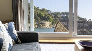 A room with a sofa, a wooden table, and a view of a coastal landscape through a large window. - South Sands Hotel (Salcombe)