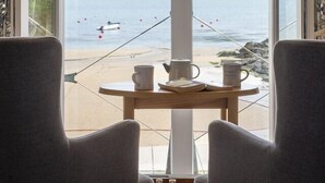 A room with a large window overlooking a beach, a table with a kettle and cups, and two chairs. - South Sands Hotel (Salcombe)