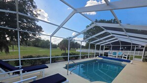 An outdoor pool area with a covered patio, lounge chairs, and a view of a grassy field. - Relaxing at the pool (Inverness)
