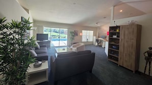 A spacious living room with a sofa, coffee table, and a TV. There is a sliding glass door leading to a balcony with a pool. - Relaxing at the pool (Inverness)