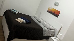 A neatly made bed with a black and grey duvet, a pillow, and folded towels. There is a bedside table with a lamp and a phone. A painting is on the wall above the bed. - Apartment in St. Peter  (Roma)