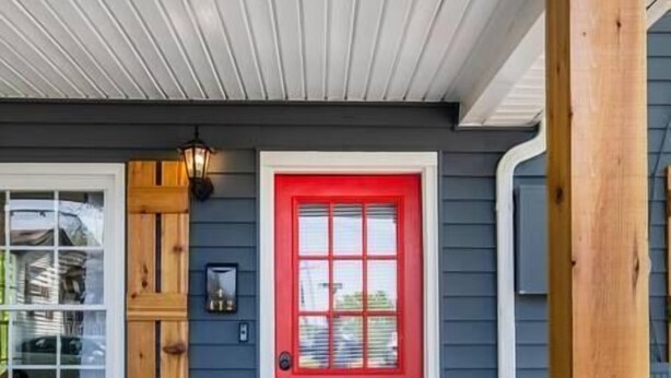 Une maison avec une porte rouge, un volet en bois et une véranda dotée d’un plancher en béton.